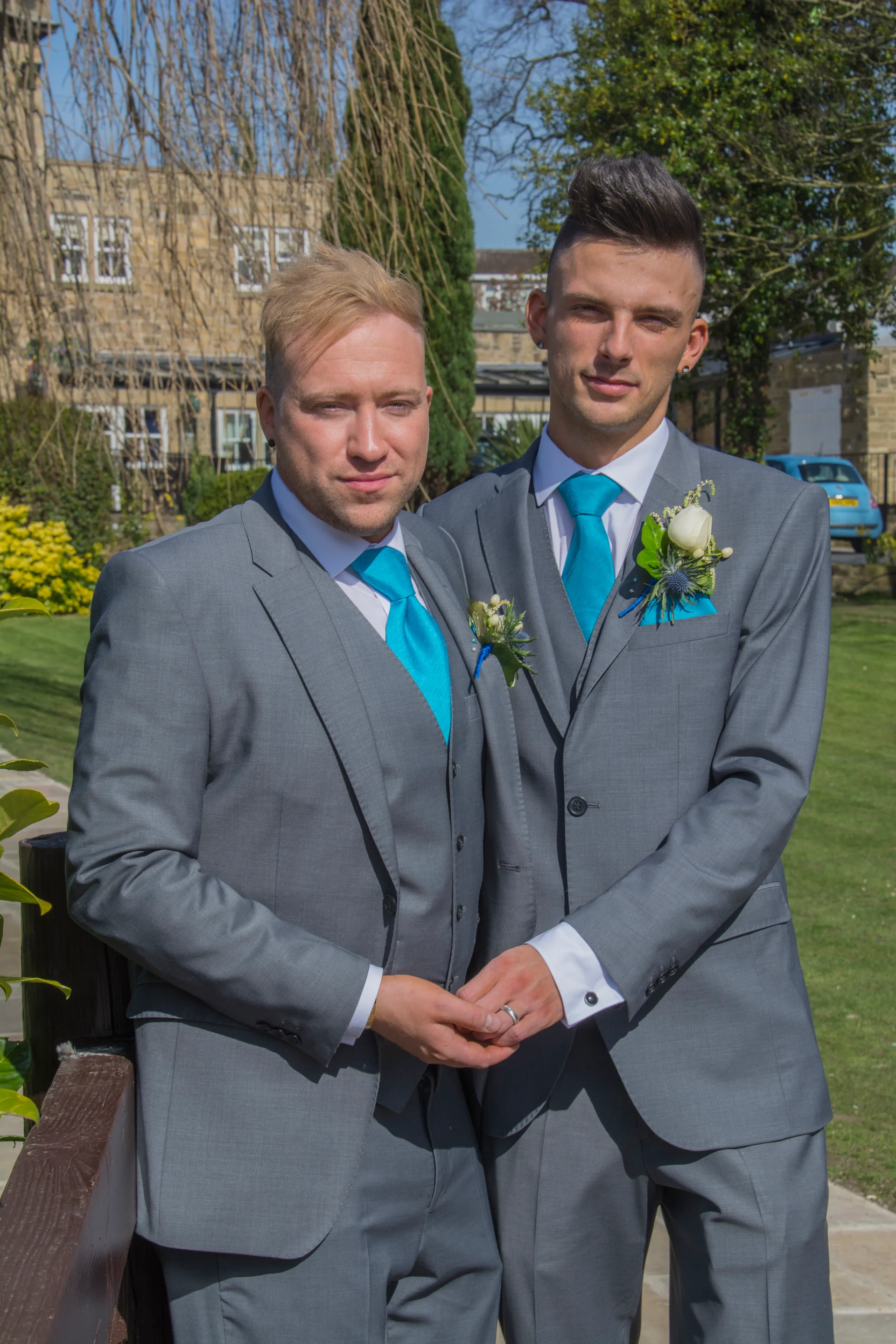 Two grooms in matching grey three-piece suits and bright blue ties hold hands outdoors on a sunny day.