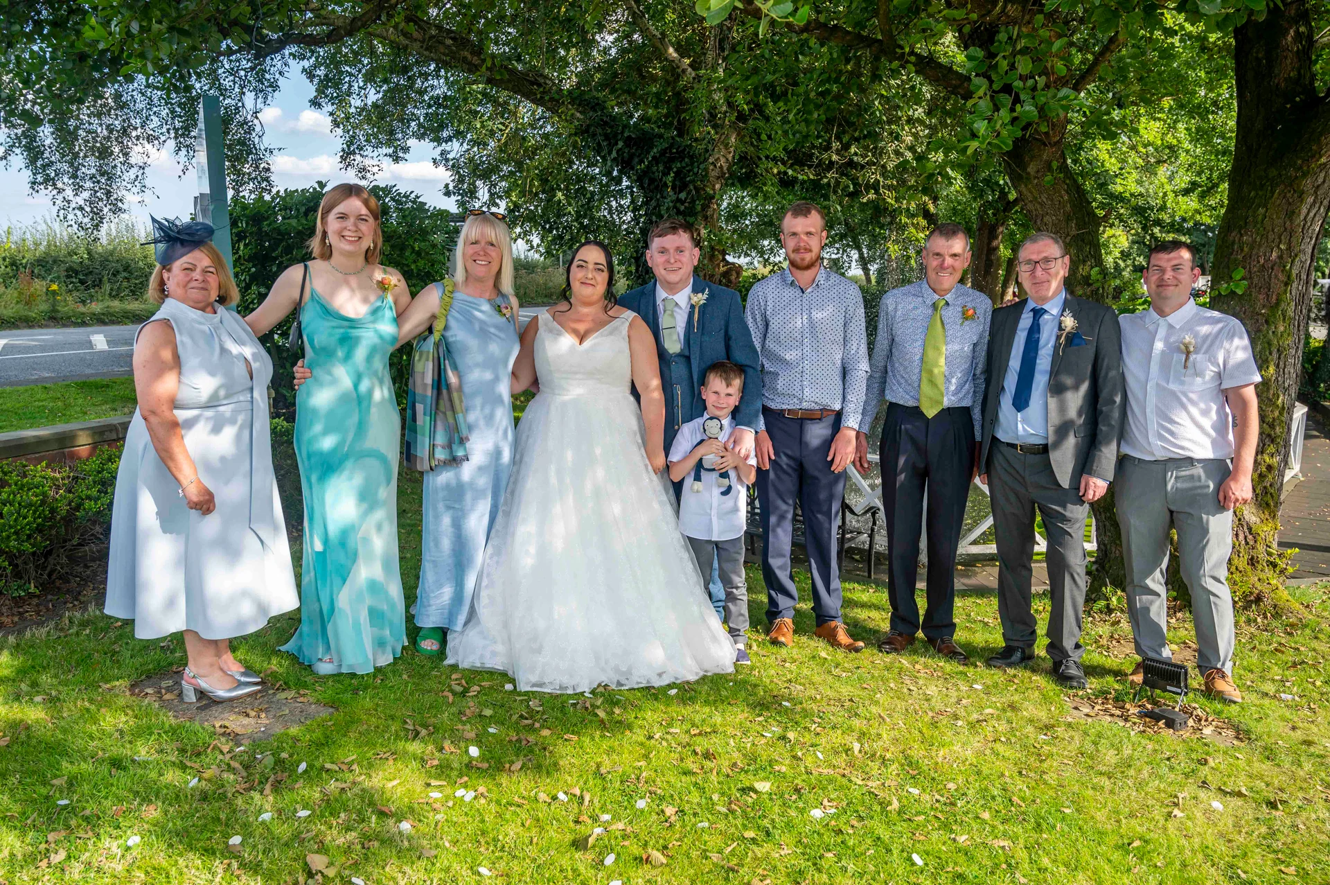 A formal family group photograph of the bride, groom, and wedding guests posing outdoors on a grassy lawn beneath the shade of mature trees on a sunny summer day.