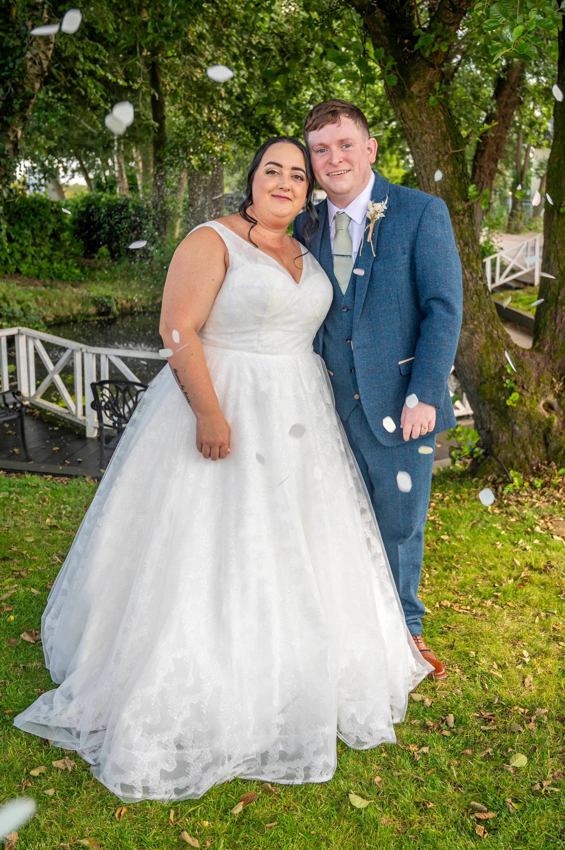 A newlywed couple stands outdoors on a lawn surrounded by falling confetti, with the bride wearing a glittery a-line gown and the groom in a blue tweed suit.