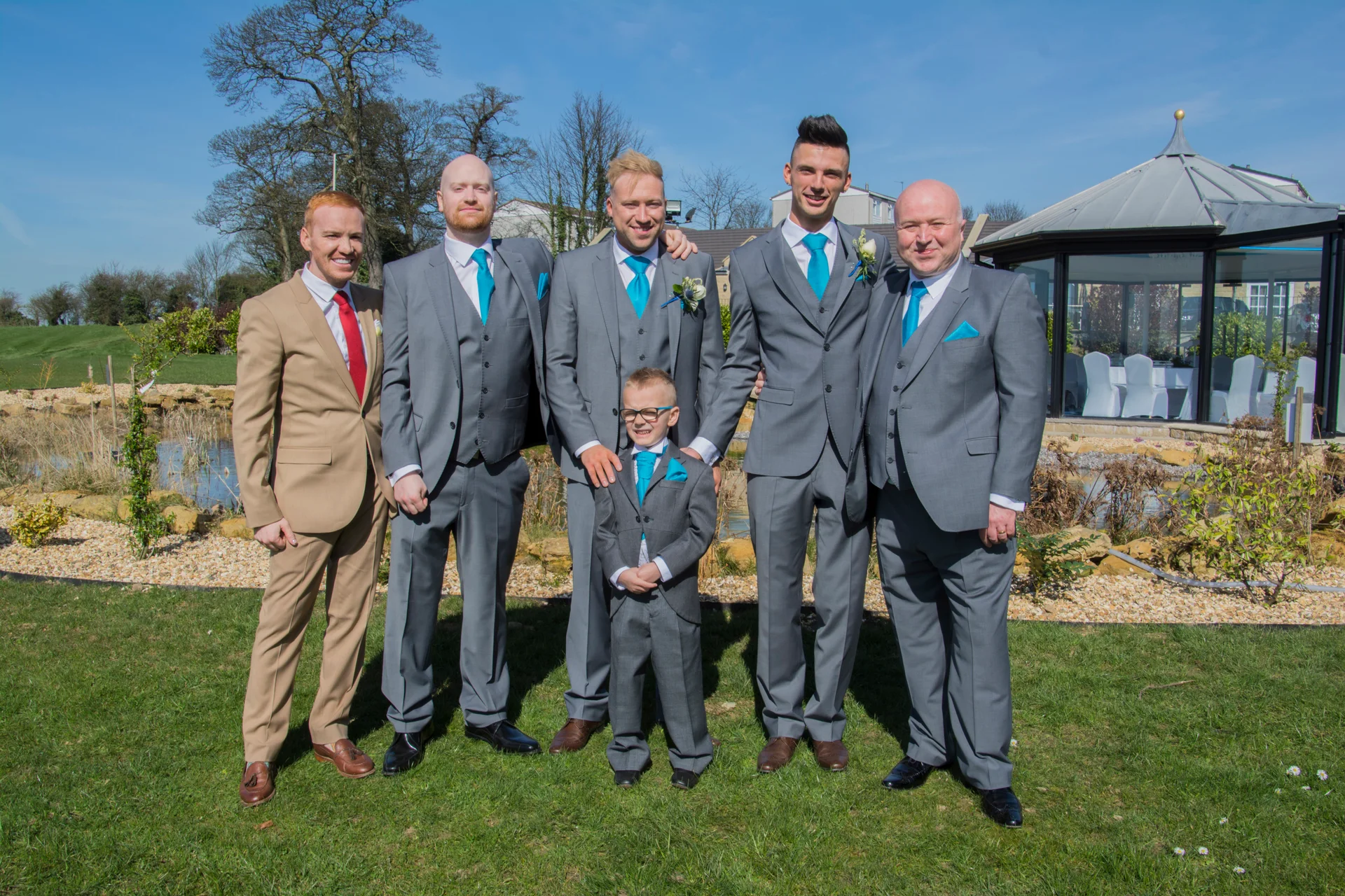 The groom and his groomsmen stand outside in matching grey three-piece suits with bright blue ties on a sunny wedding day.