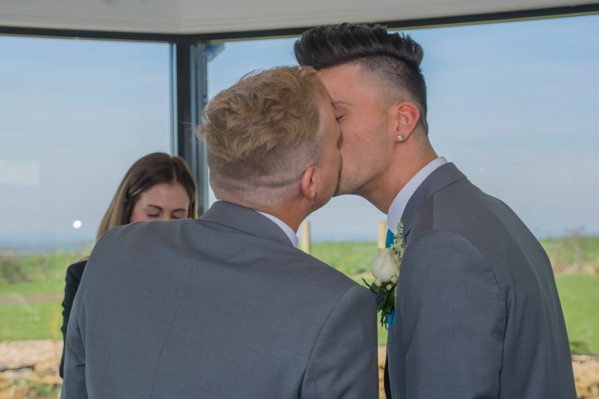 Two grooms share their first kiss as a married couple during a bright indoor ceremony wearing matching grey suits.