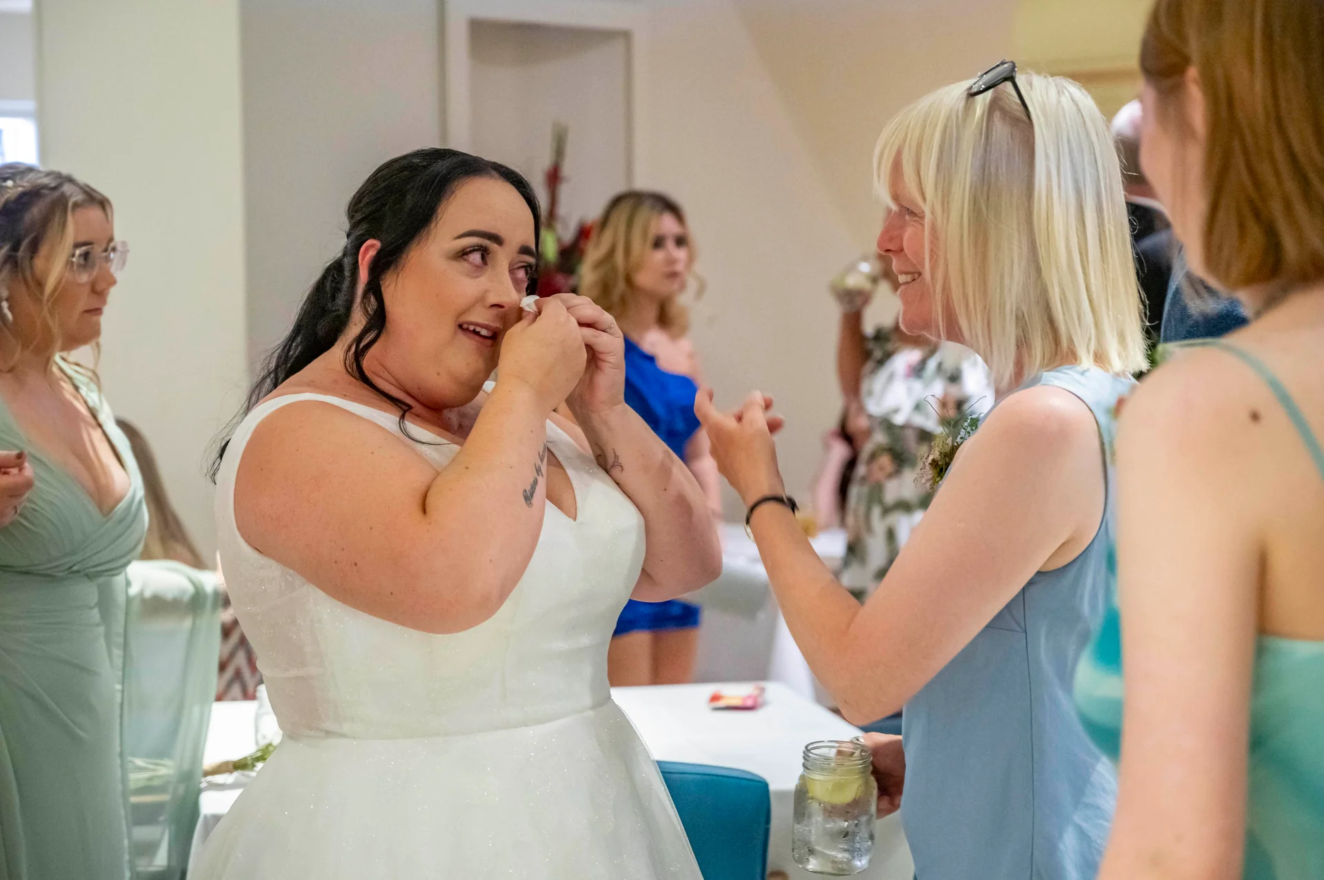 An emotional bride in a sparkly white wedding dress wipes a tear away with a tissue while chatting with guests during the reception.