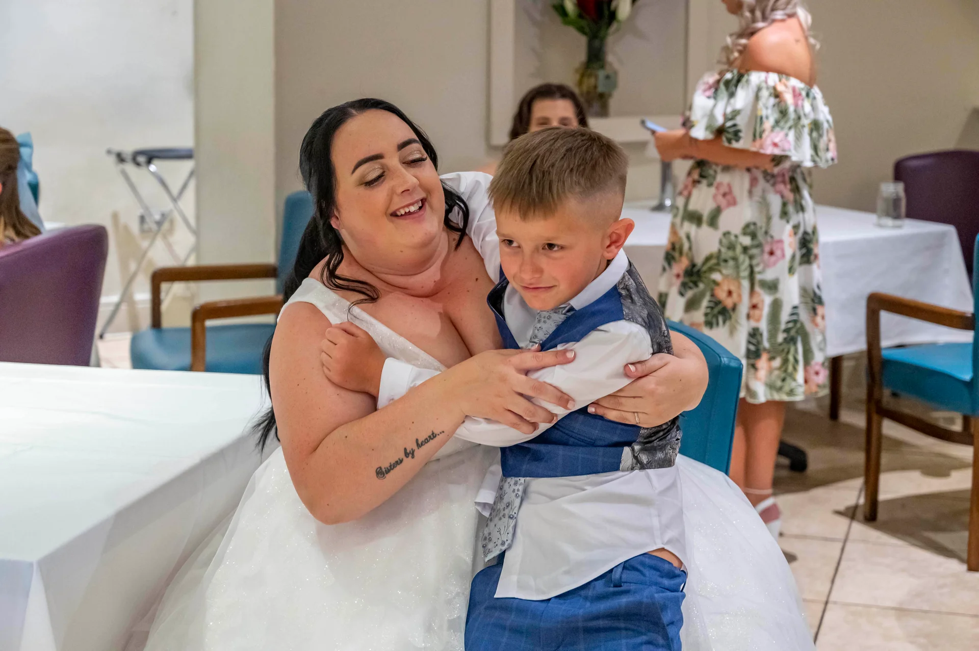 A smiling bride in a white tulle gown warmly hugs a young page boy wearing a blue patterned waistcoat at a wedding reception.