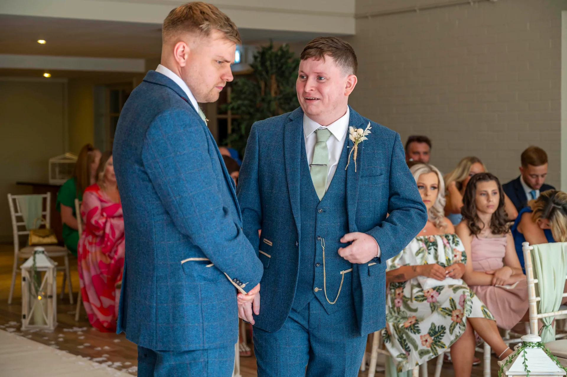 Two grooms in matching blue tweed suits stand together at the altar during their wedding ceremony while guests watch from seated rows.