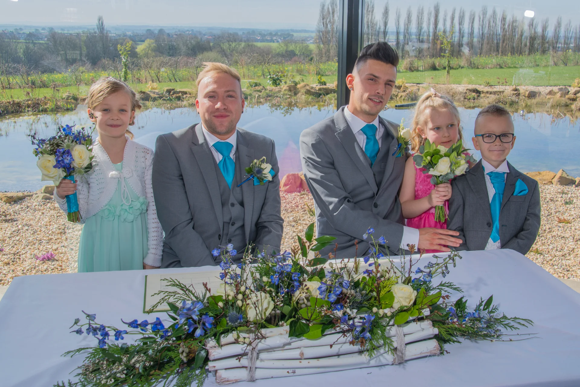 Two grooms in matching grey suits and bright blue ties sit together at a registry table decorated with white and blue floral arrangements, accompanied by young flower girls and a page boy.