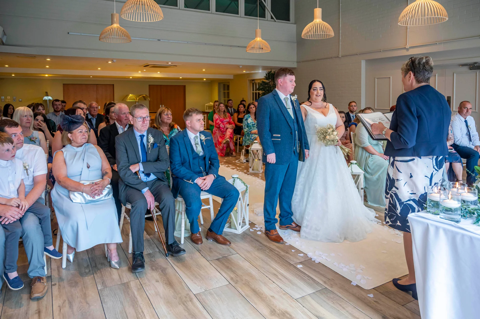 A bride and groom stand at the altar during a civil wedding ceremony while guests watch attentively from seated rows.