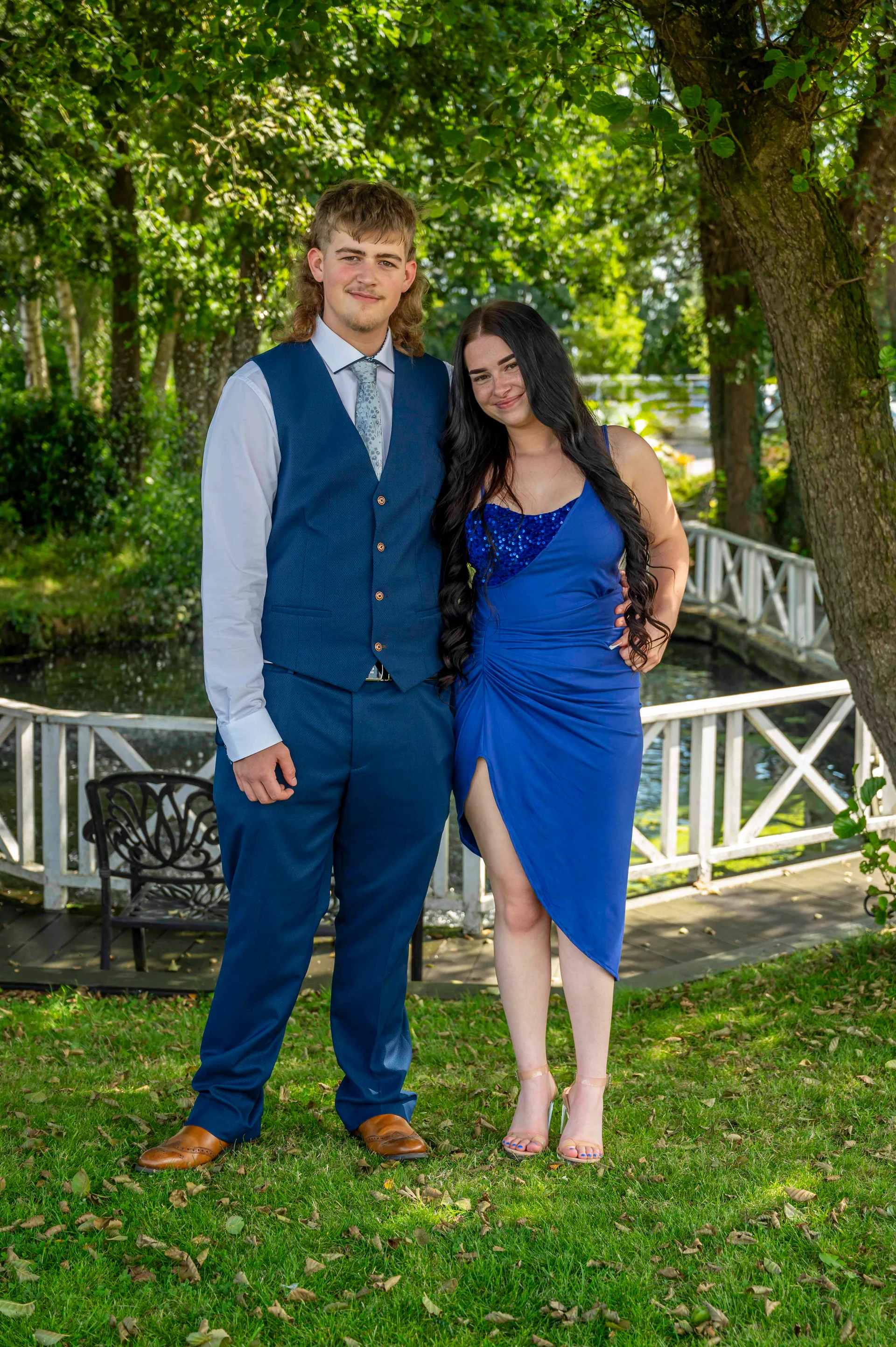 A young couple poses outdoors on a grassy lawn near a lake and a white wooden bridge, wearing a blue waistcoat and a royal blue dress.