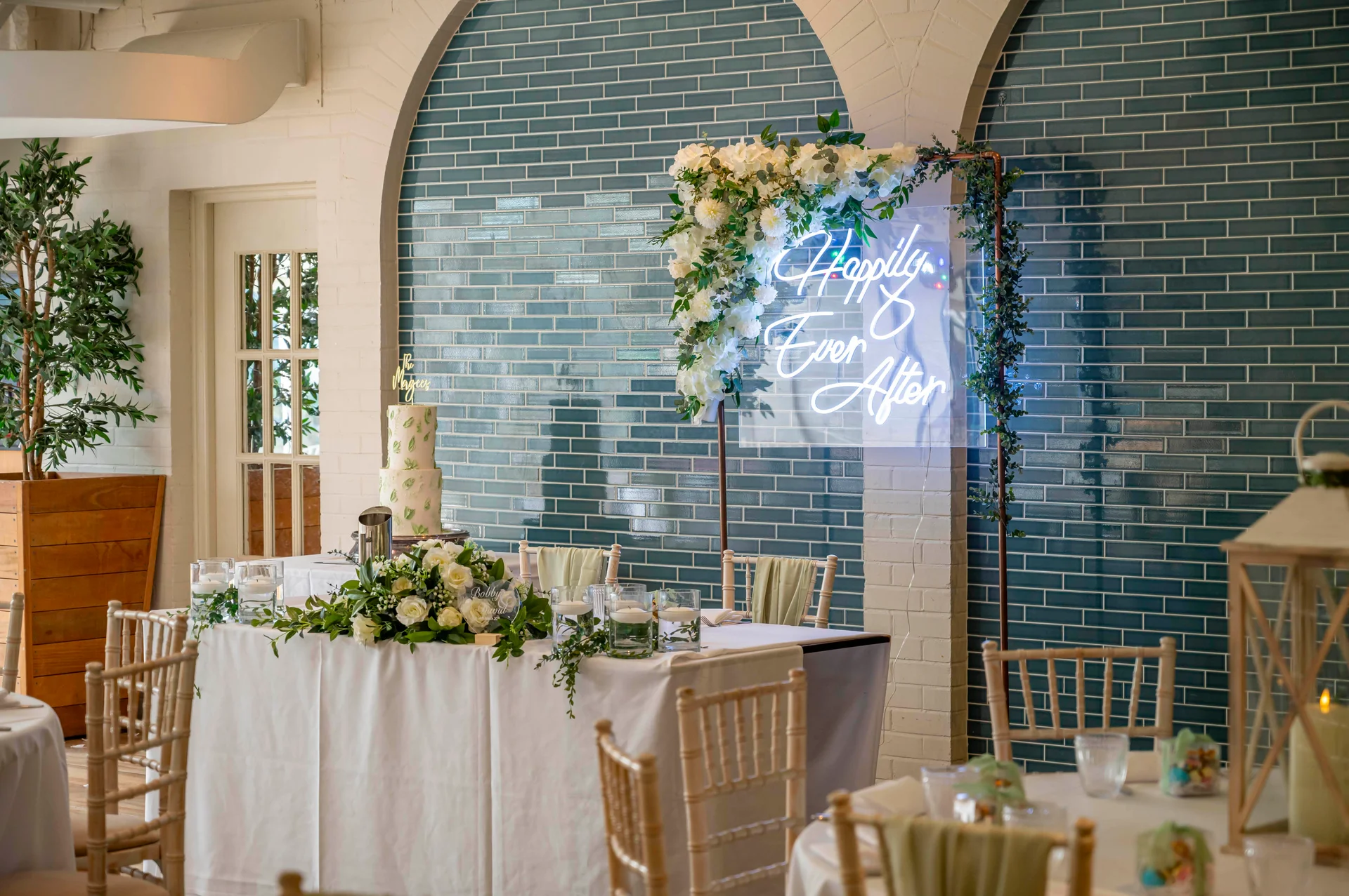 A beautifully styled wedding top table features white floral arrangements, a two-tier cake, and a glowing neon sign against a blue-tiled industrial backdrop.