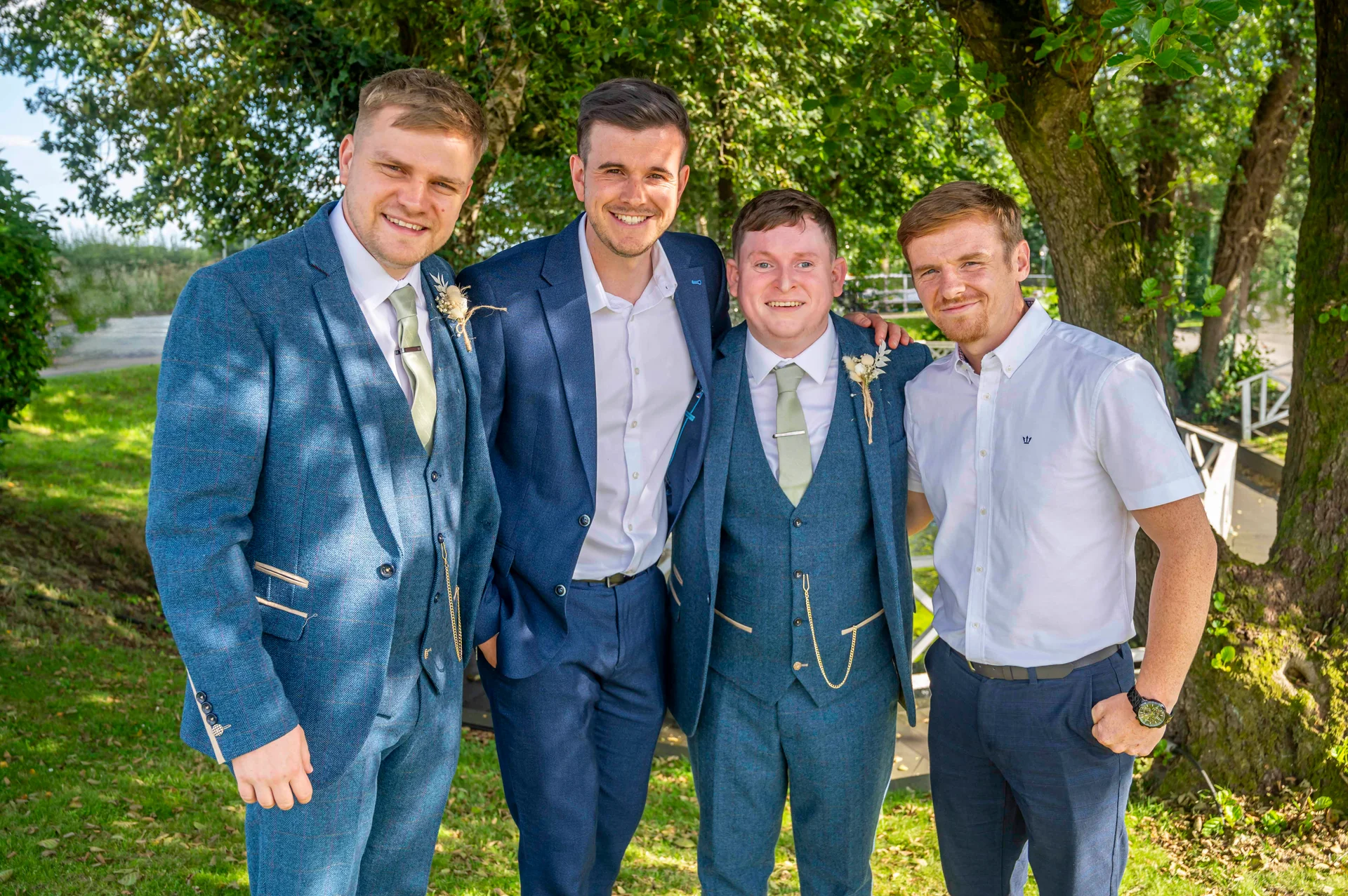 Four smiling groomsmen in blue tweed suits and open shirts stand outdoors beneath the dappled shade of leafy trees.