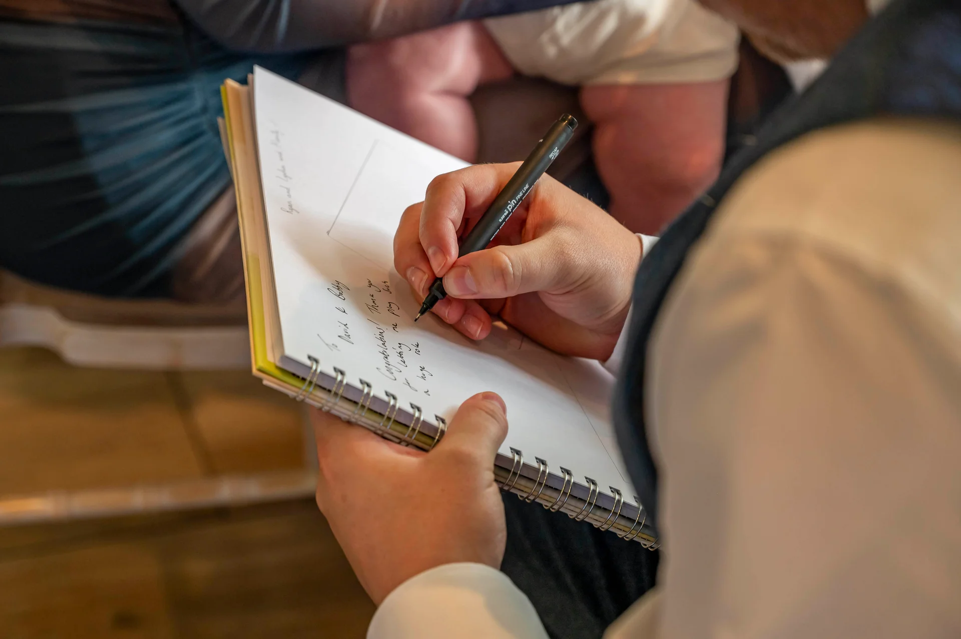 A close-up over-the-shoulder shot captures a guest writing a heartfelt message in a spiral-bound wedding guest book using a black pen.