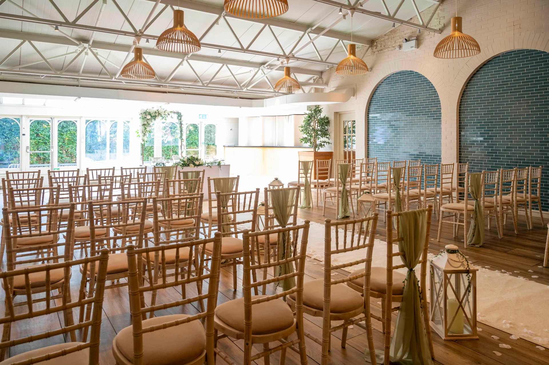 A bright, modern industrial wedding venue set up for a ceremony with wooden chiavari chairs, sage green fabric drapes, lanterns along the aisle, and exposed metal roof trusses.