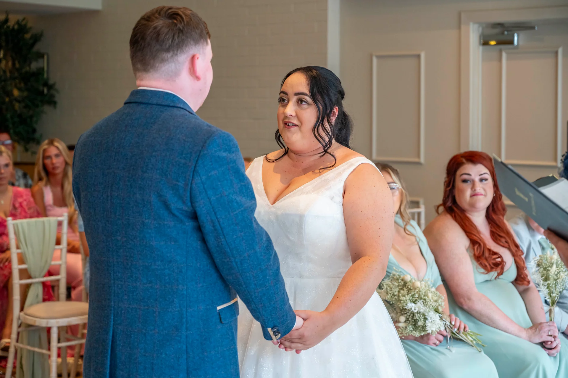 A groom in a blue tweed suit and a bride in a sparkling v-neckline gown hold hands during their indoor civil ceremony while seated guests look on.