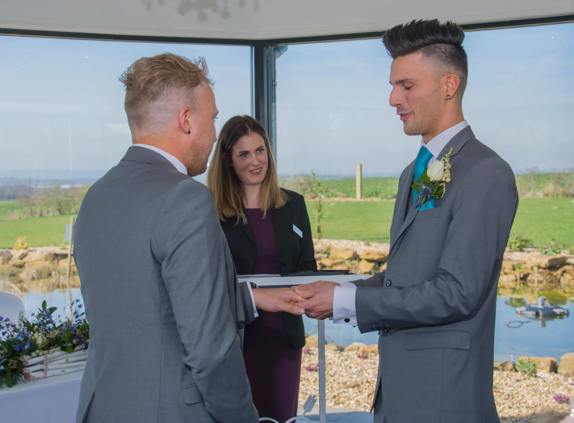 Two grooms in matching grey suits exchange rings during their outdoor civil ceremony while a celebrant looks on.
