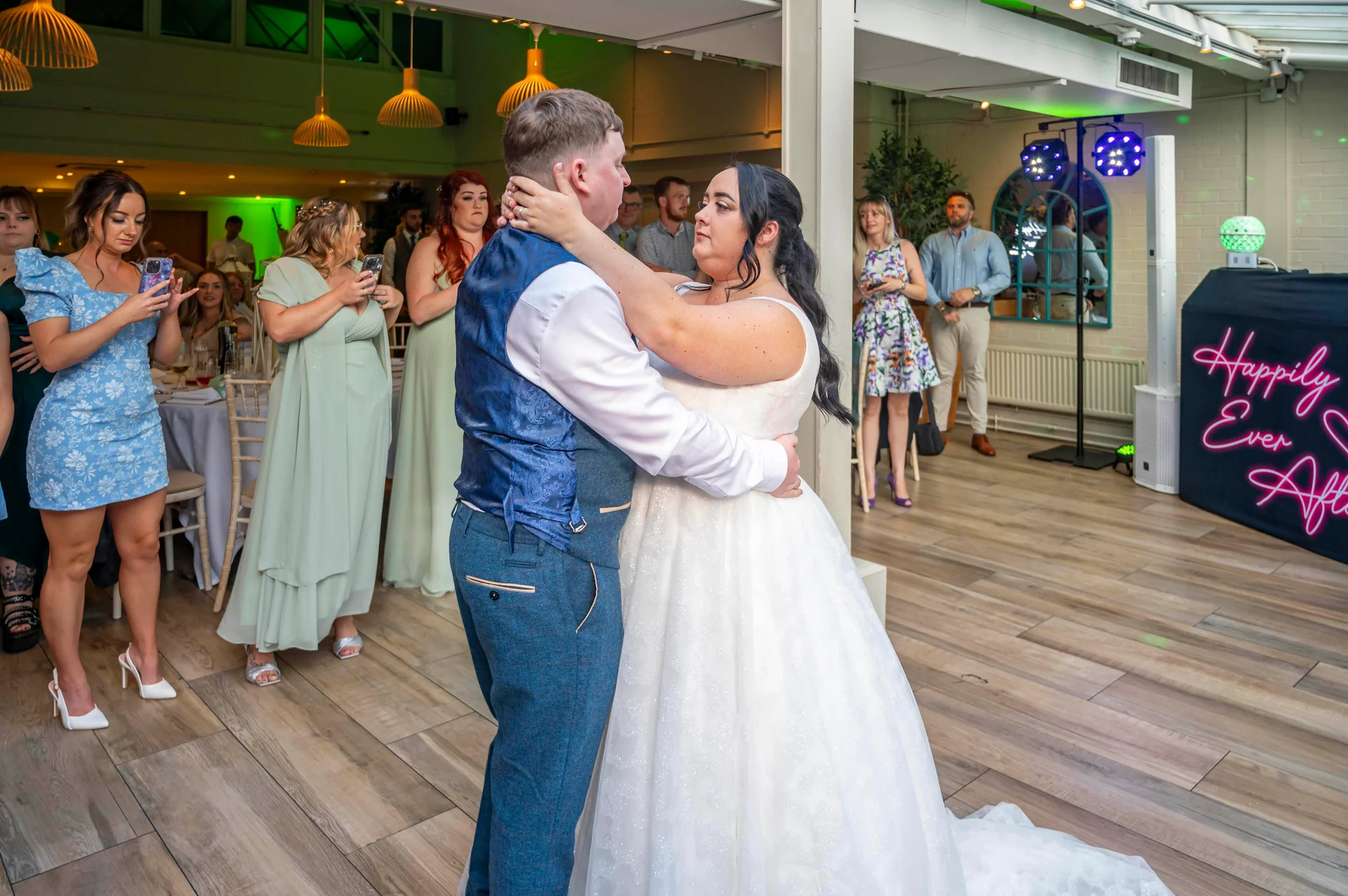 Newlyweds share an intimate embrace during their first dance on the wooden floor of a reception venue illuminated by disco lights and a glowing neon sign.