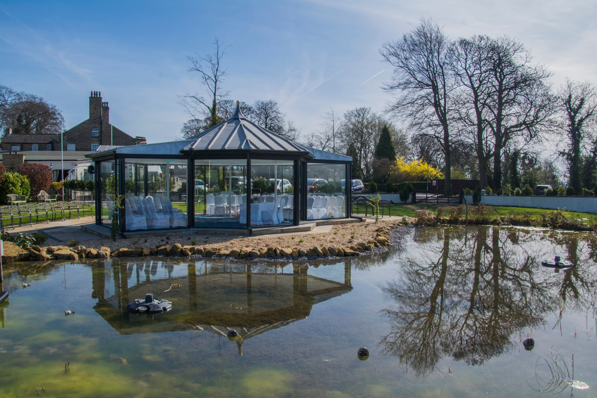A modern glass gazebo wedding venue sits beside a serene pond reflecting its structure and surrounding trees under a bright blue sky.