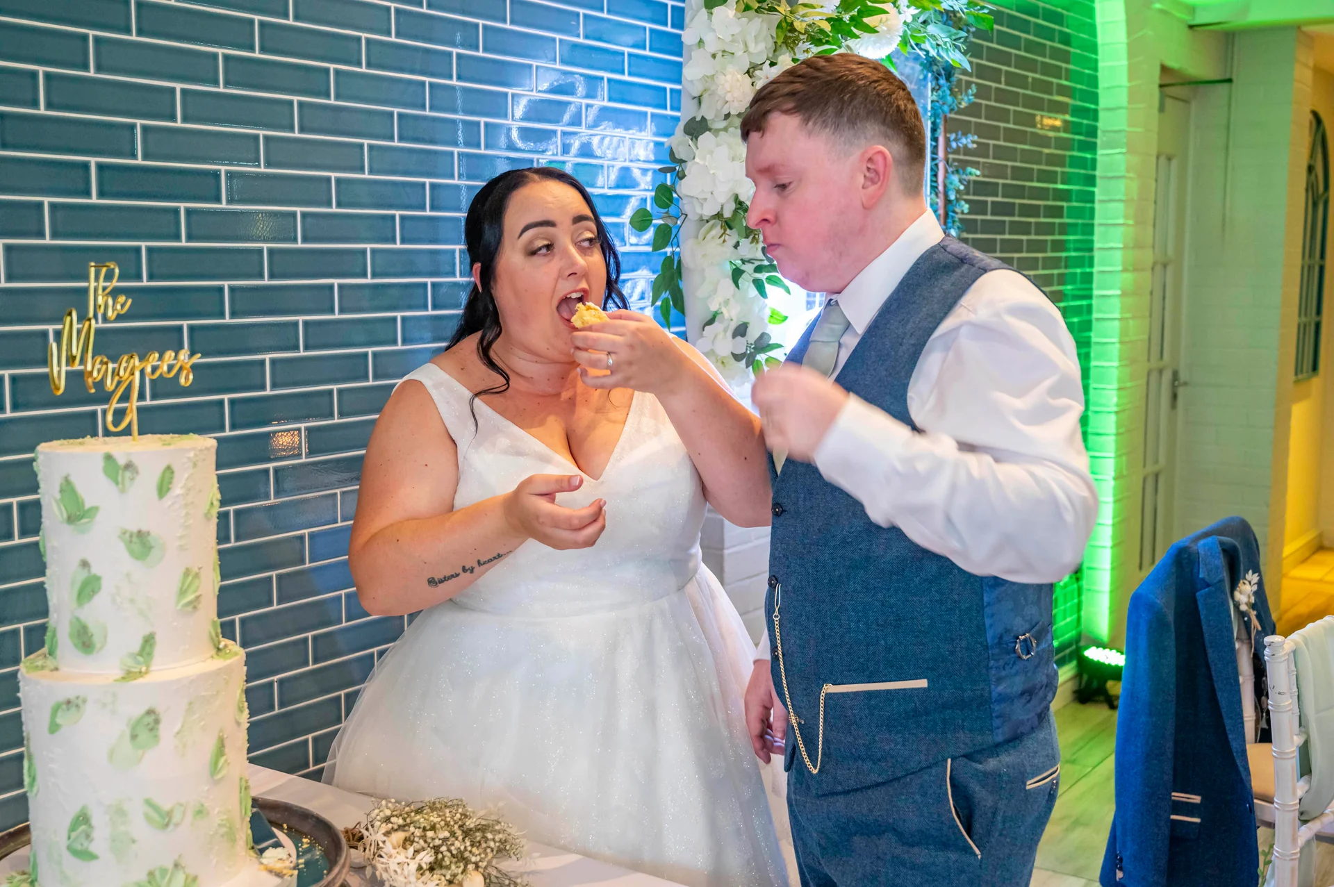 A joyful bride and groom share a playful moment feeding each other cake in front of a blue tiled wall at their evening reception.