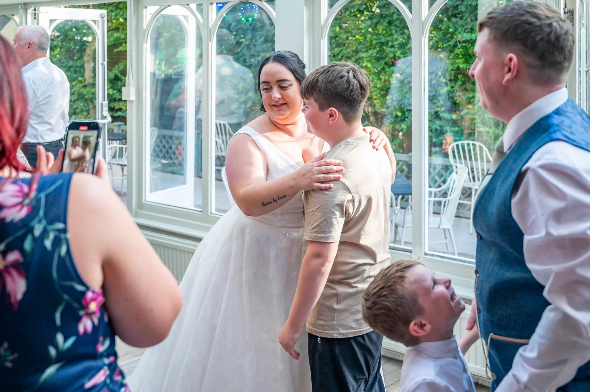 A joyful bride in a white a-line wedding dress embraces a young guest inside a bright conservatory while another child looks up and a guest photographs the moment on a mobile phone.