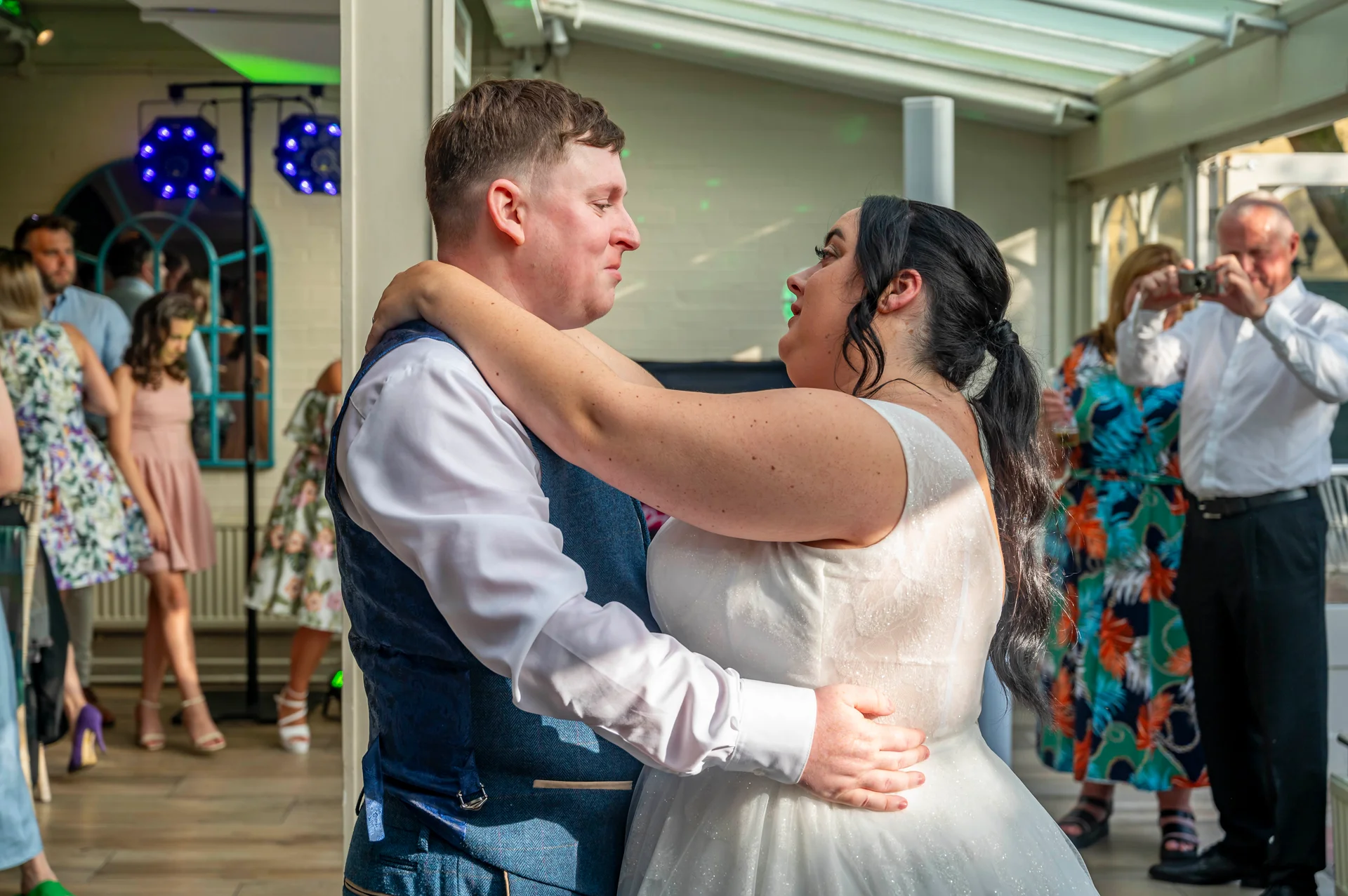 The newly married couple share a romantic first dance together inside a bright conservatory while guests look on and capture the moment.