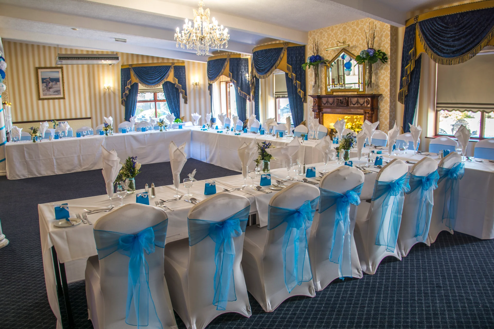 An elegantly set U-shaped wedding reception banquet table features crisp white linens, matching chair covers adorned with vibrant blue sashes, and refined glassware under a classic crystal chandelier.