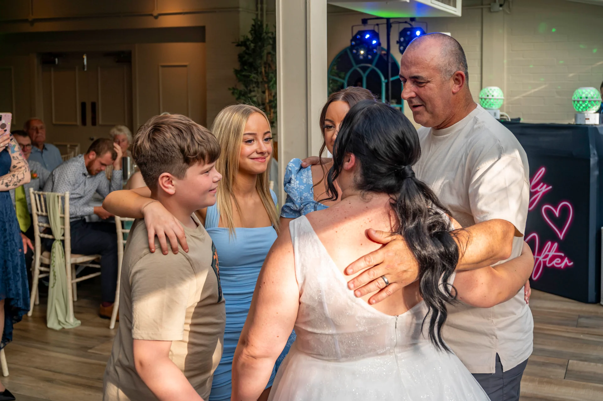 A bride in a sparkly white wedding dress embraces guests during a joyful celebration at an evening reception.