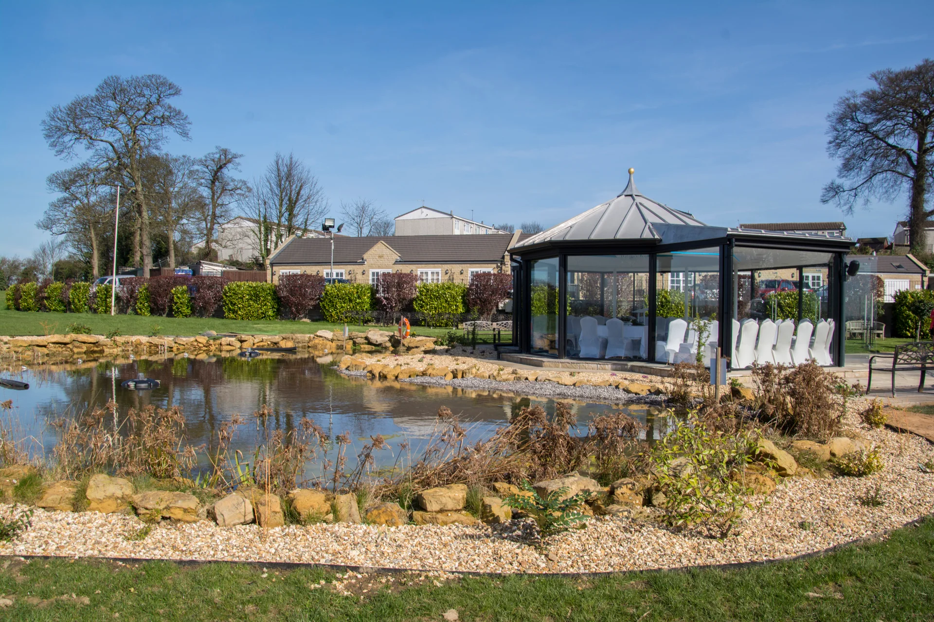 A modern glass gazebo wedding venue stands beside a tranquil lake under a bright blue spring sky.