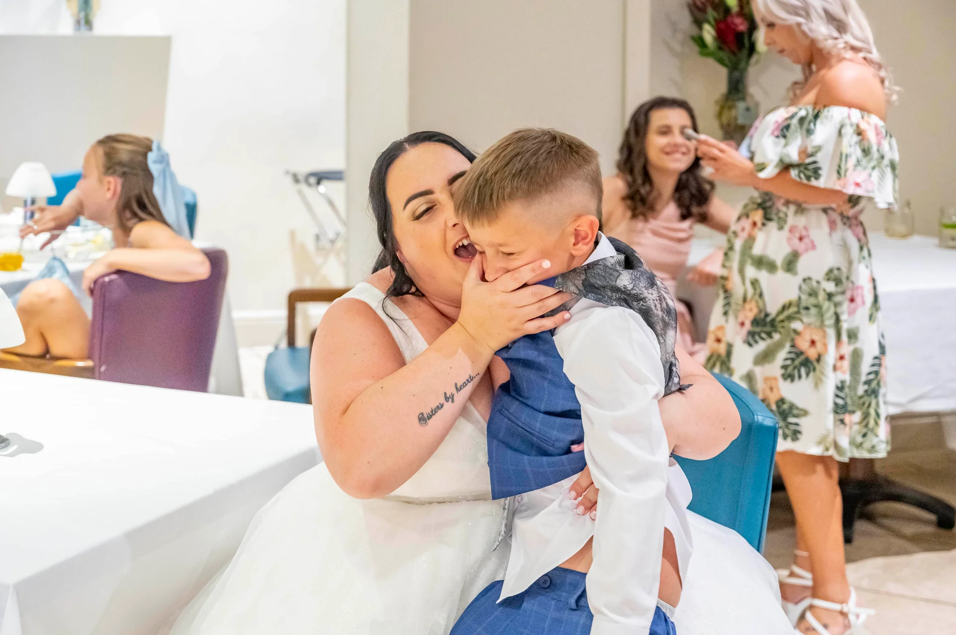 A joyous bride sharing a tender, laughing embrace with a young page boy in a blue suit during the wedding reception.
