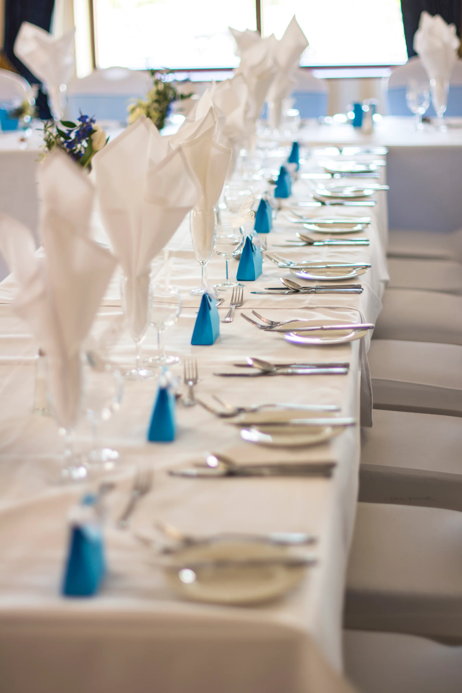 A long wedding banquet table is meticulously set with white linens, tall folded napkins, and vibrant blue favor boxes under bright, natural light.