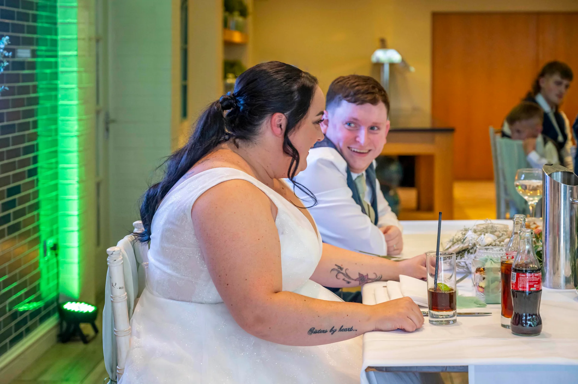 The bride laughs heartily at the wedding breakfast while the groom looks on lovingly from across the top table.