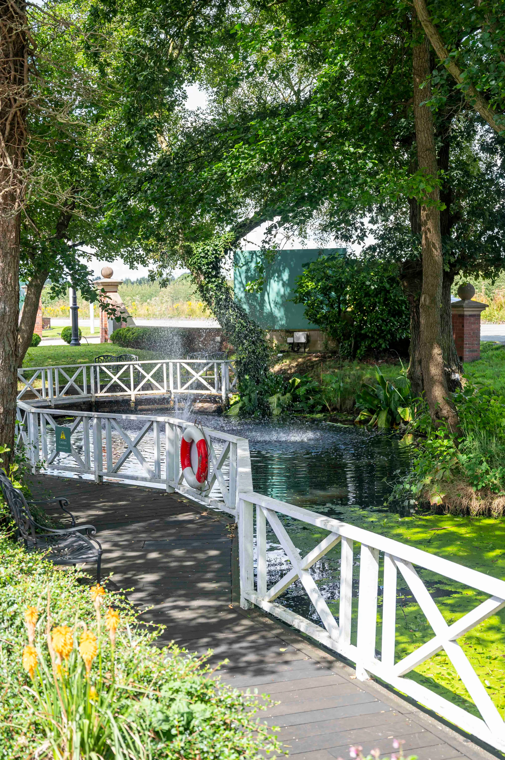 A scenic garden pathway features a white wooden bridge over a pond with a fountain, surrounded by lush green trees and foliage.