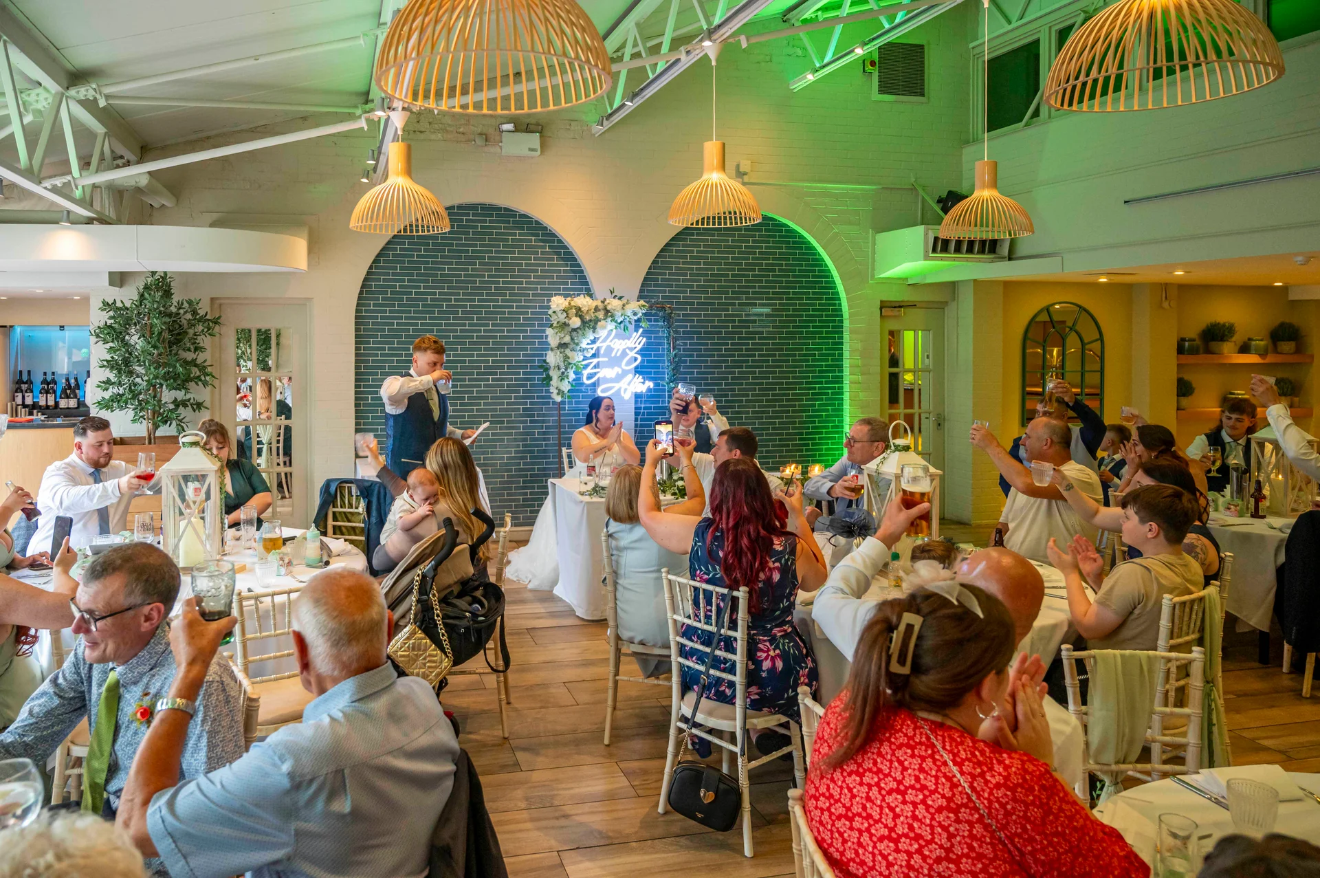 Guests raise their glasses and applaud during the speeches at a wedding reception set in a modern industrial venue featuring distinctive wooden pendant lighting and teal tiled walls.