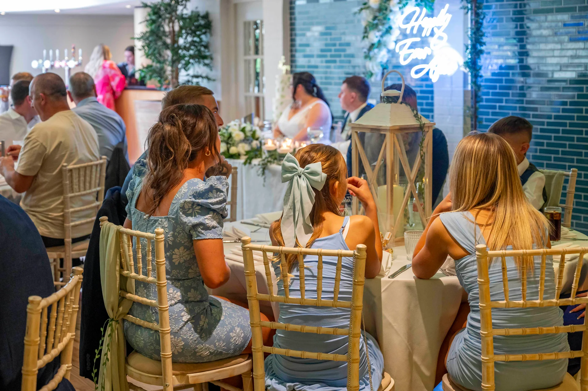 Guests sit at a round table during the wedding breakfast, viewed from behind with blue dresses and a decorative lantern on the table.