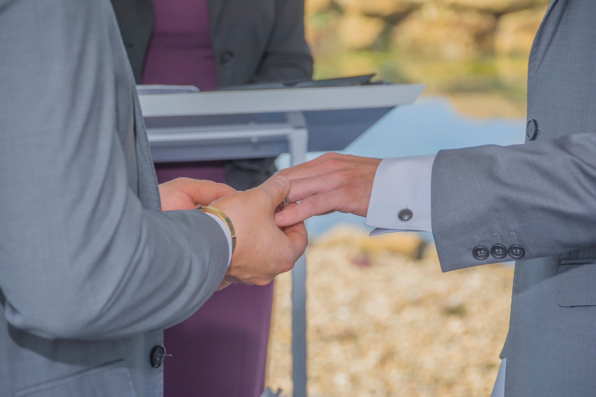 Two grooms in matching grey suits exchange wedding bands during an outdoor ceremony, with a close-up on their hands as the ring is placed.