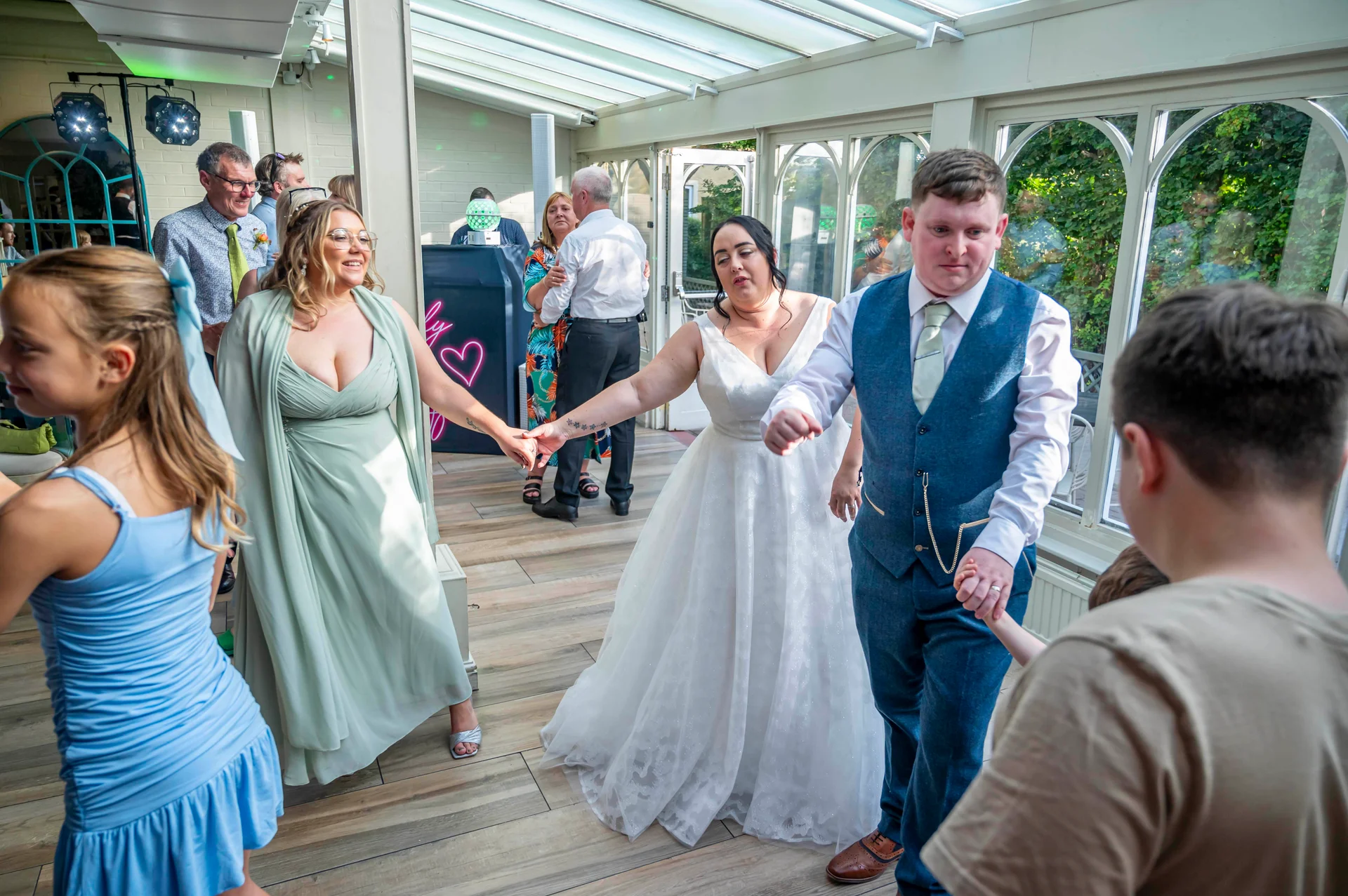 Newlyweds and guests hold hands while dancing on the wooden floor inside a bright conservatory during the evening reception.