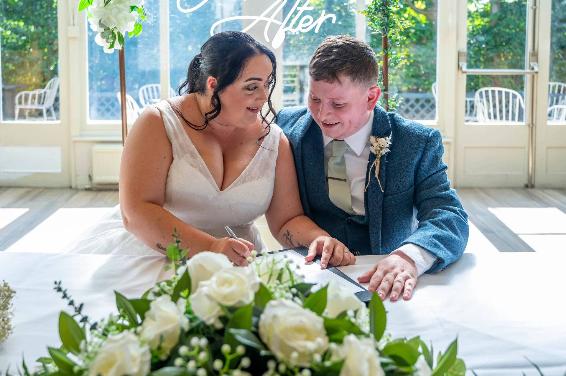 A smiling bride and groom sign the register together in a light-filled orangery decorated with white roses.