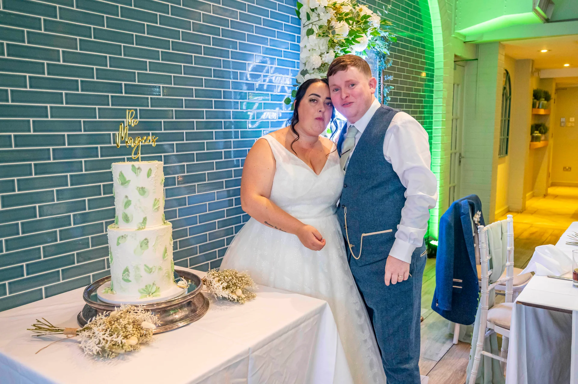A newlywed couple poses next to a two-tier wedding cake in front of a blue tiled wall and green ambient lighting.