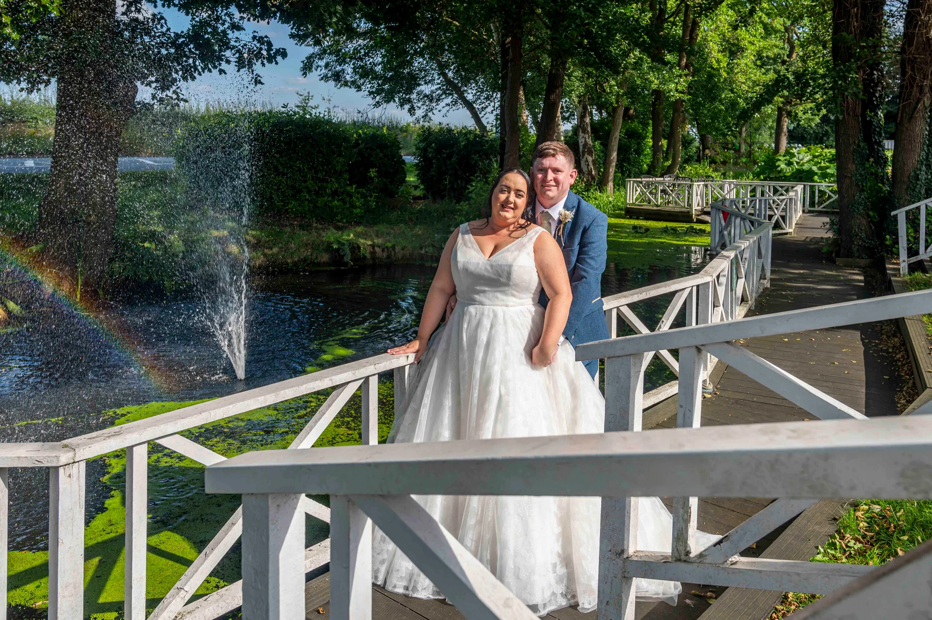 A bride in a white a-line gown and groom in a blue suit pose on a white wooden bridge over a lake, with a fountain and rainbow in the background.