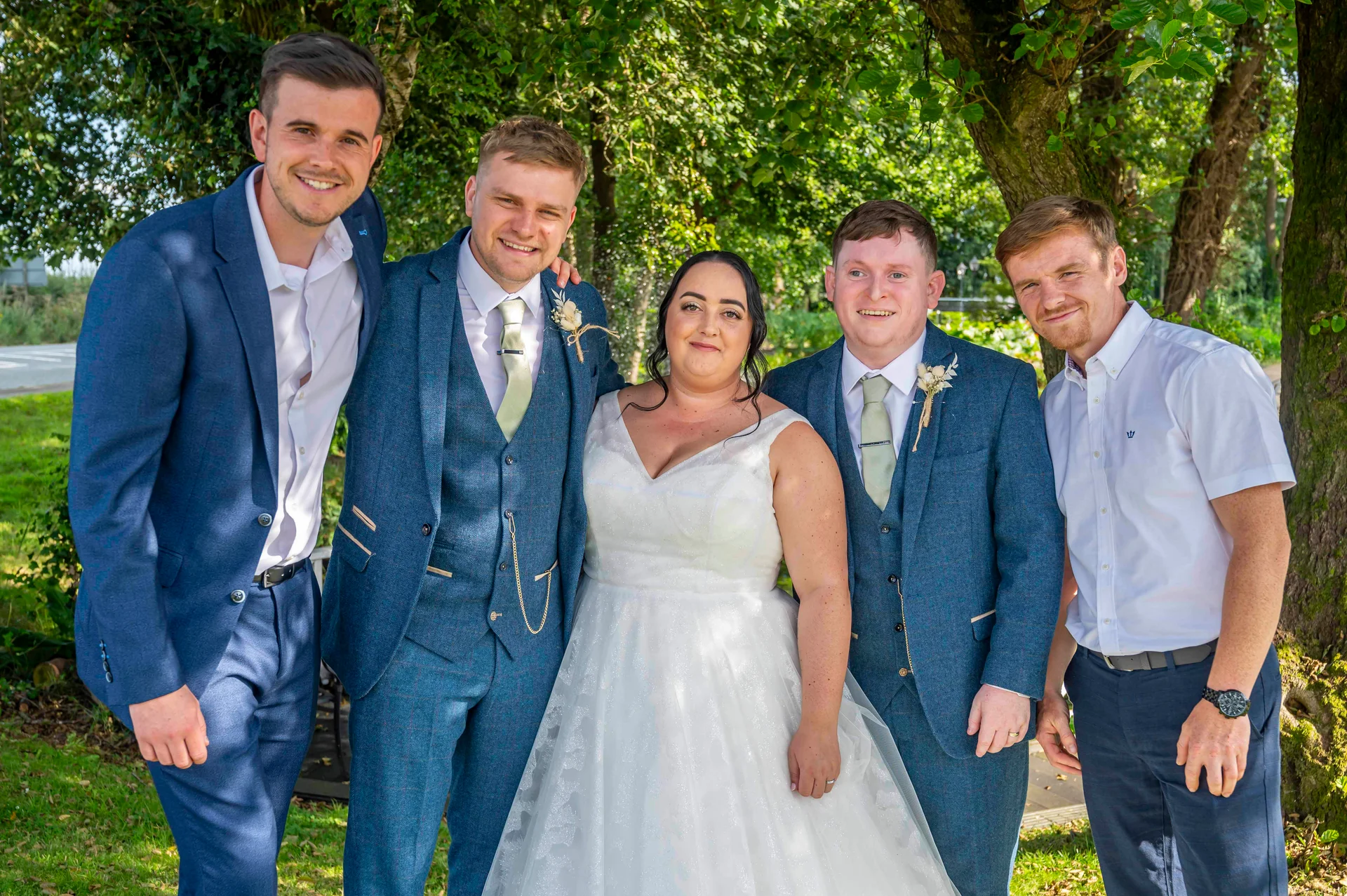 The bride in an a-line white gown stands proudly among the groom and groomsmen wearing blue tweed suits and matching sage green ties outdoors in a sunlit garden.