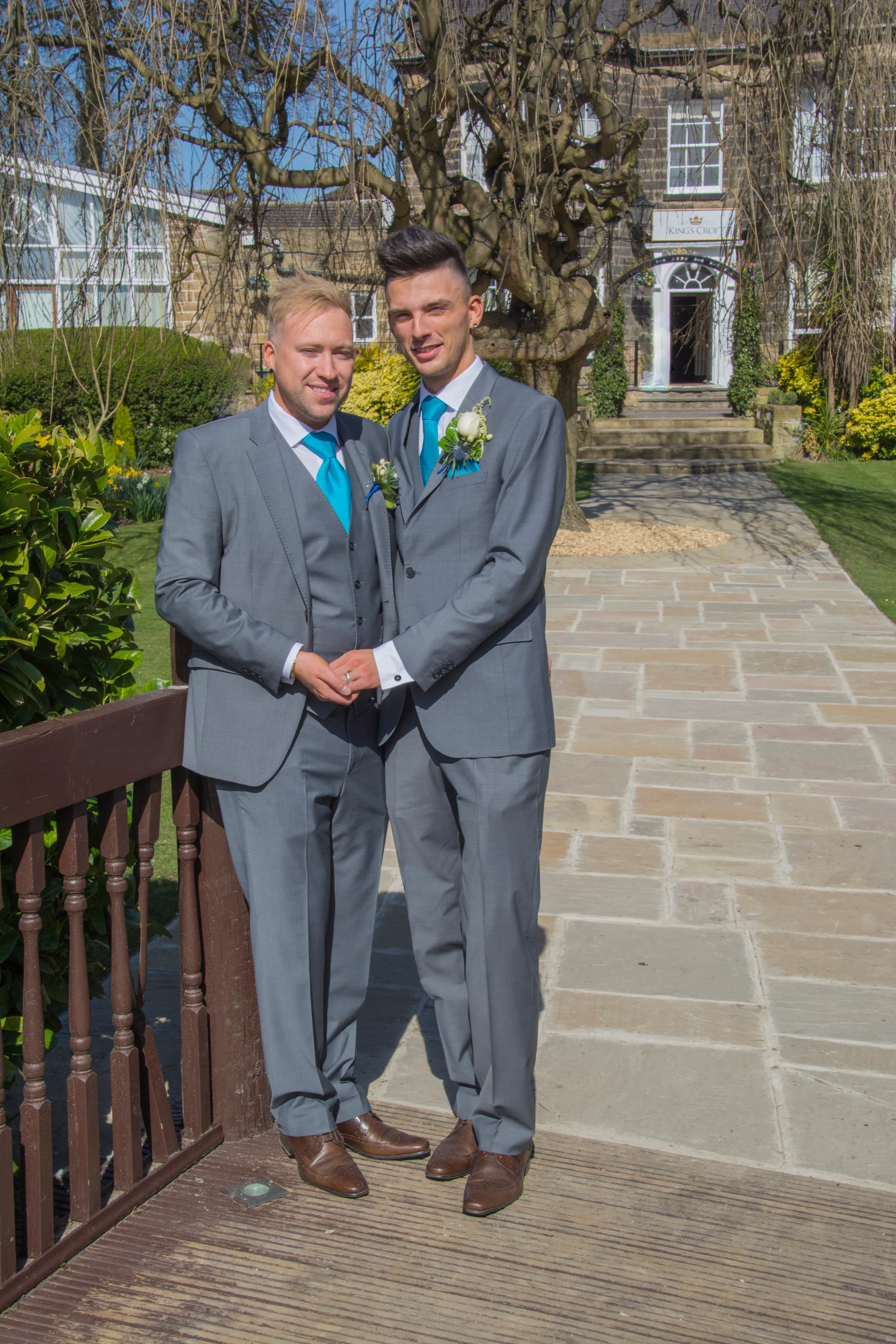 Two grooms in matching grey suits and bright blue ties stand together holding hands on a stone bridge outside a manor house.