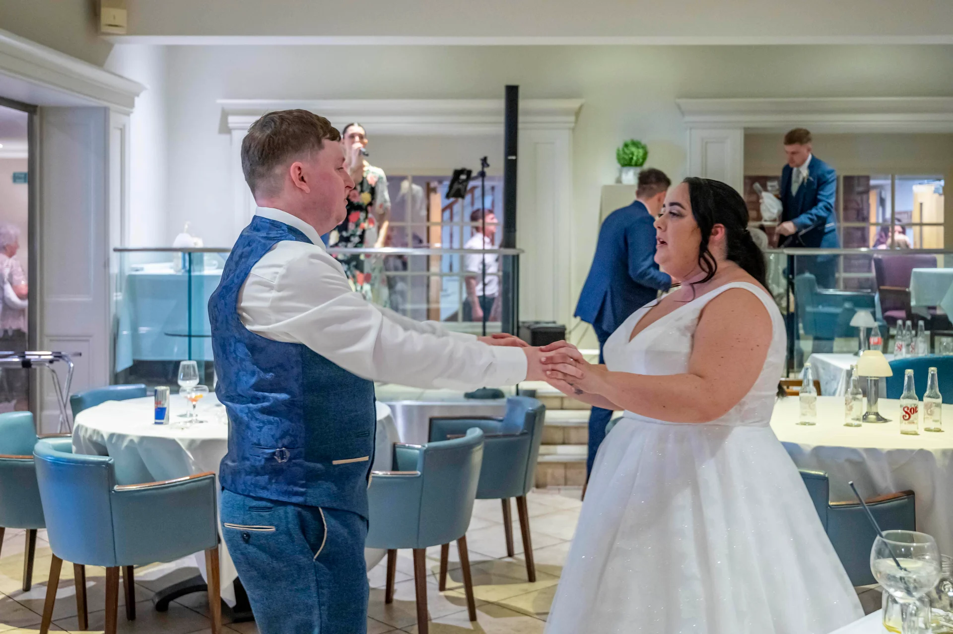 A newlywed couple share their first dance together in a hotel reception venue with guests mingling in the background.