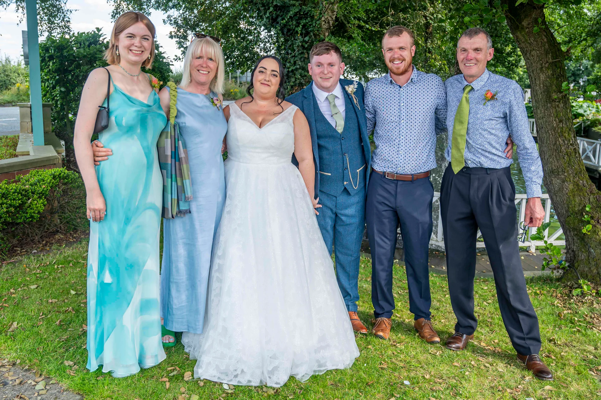 A happy wedding party poses outdoors on a grassy lawn beside a lake, featuring the bride in a white gown and the groom in a tweed suit alongside family members in blue attire.