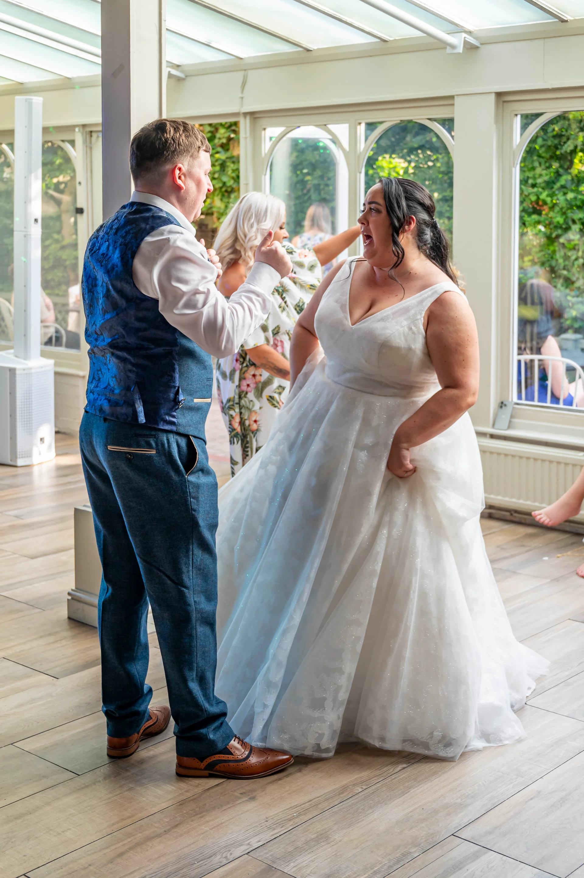 A newlywed couple joyfully dances together inside a bright conservatory with large arched windows during their wedding reception.