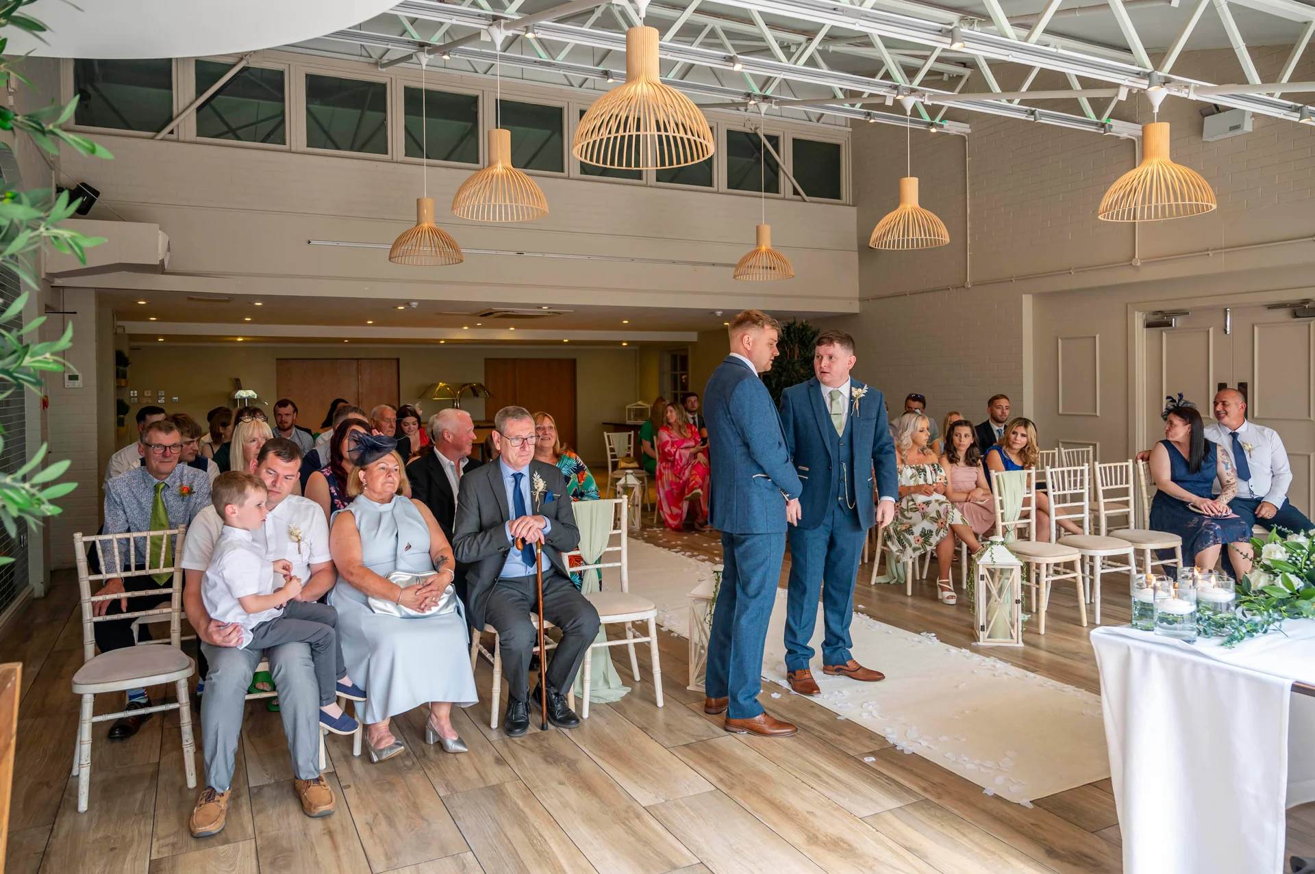 Two grooms stand at the front of a bright, modern ceremony room while seated guests watch on during their wedding.