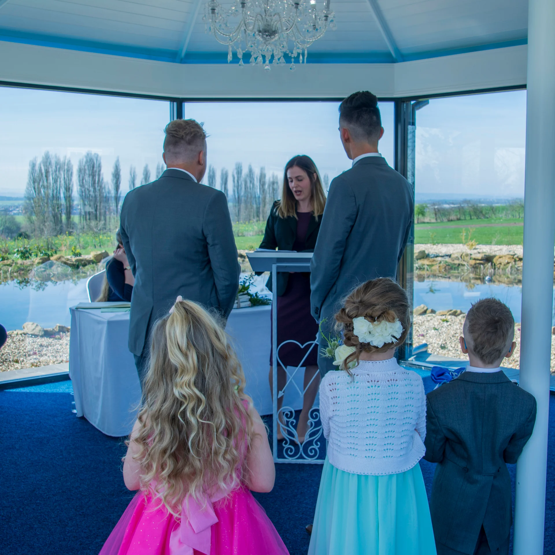 Two grooms stand at the altar during a light-filled civil ceremony overlooking a serene lakeside and rolling hills, viewed from behind with children in the foreground.