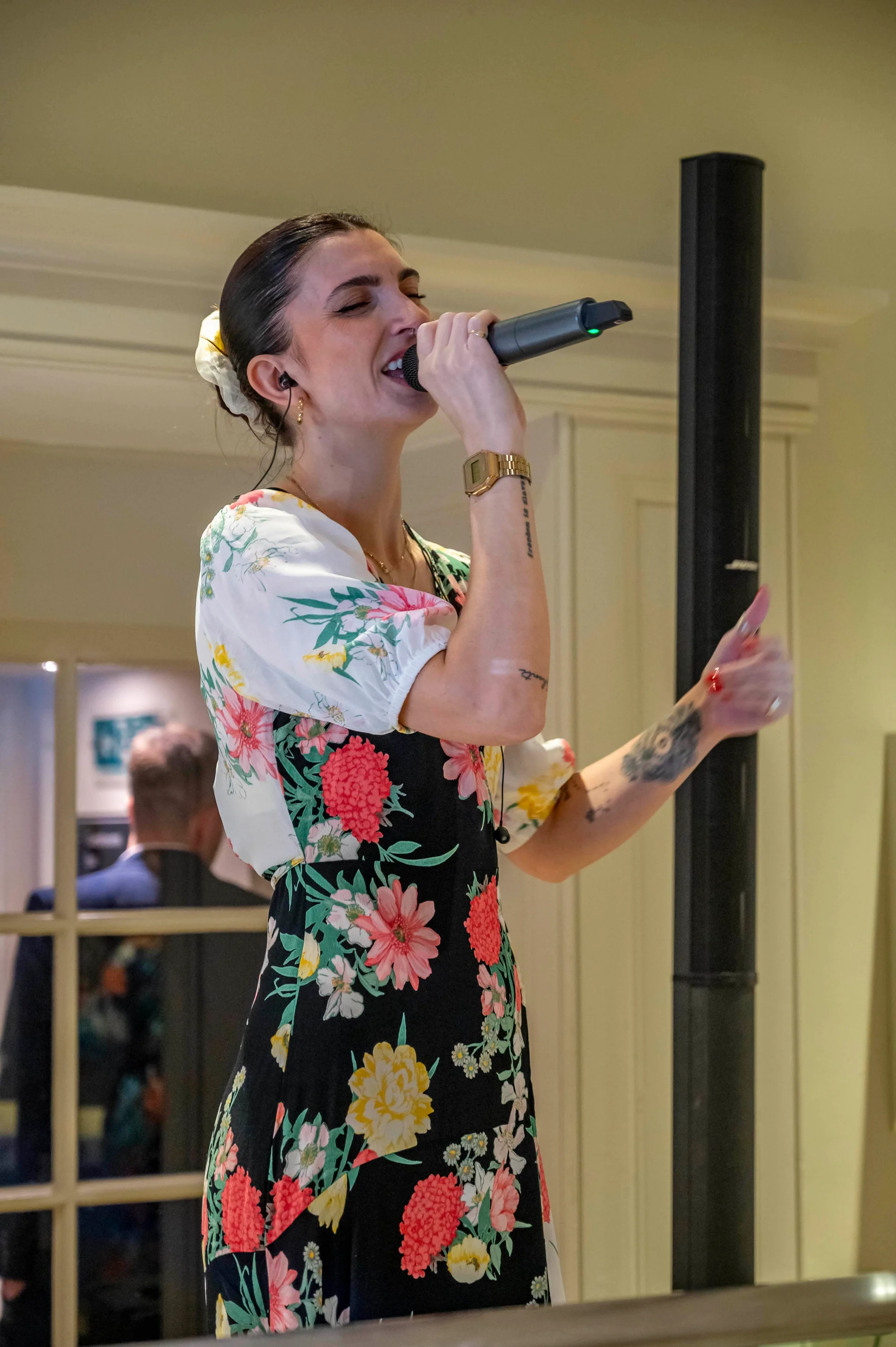 A female singer with dark hair tied back in a floral dress performs enthusiastically into a microphone during a wedding celebration.
