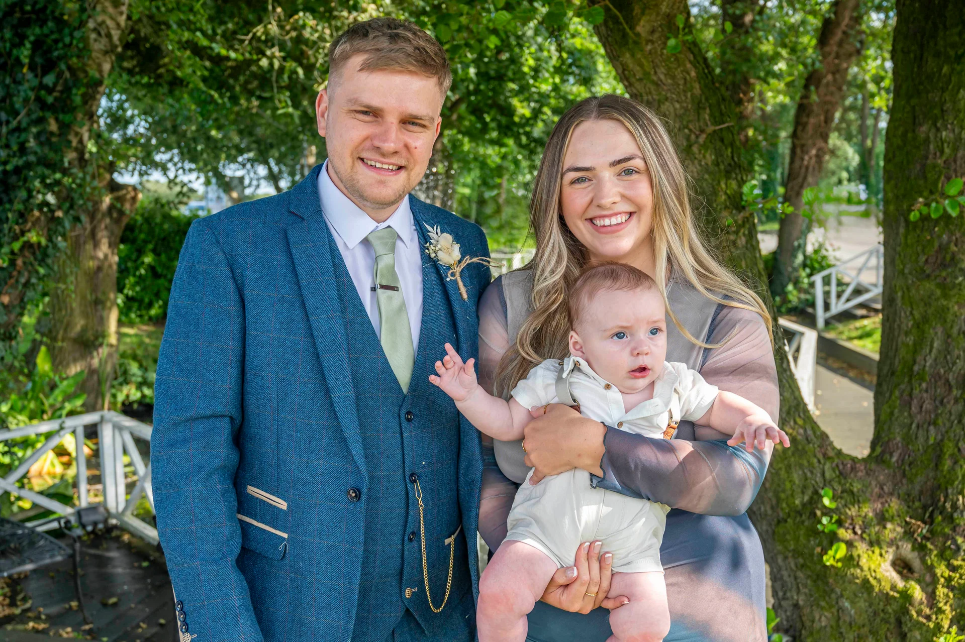 A smiling couple poses outdoors in an outdoor setting, with the groom wearing a blue three-piece suit and the bride holding a baby.