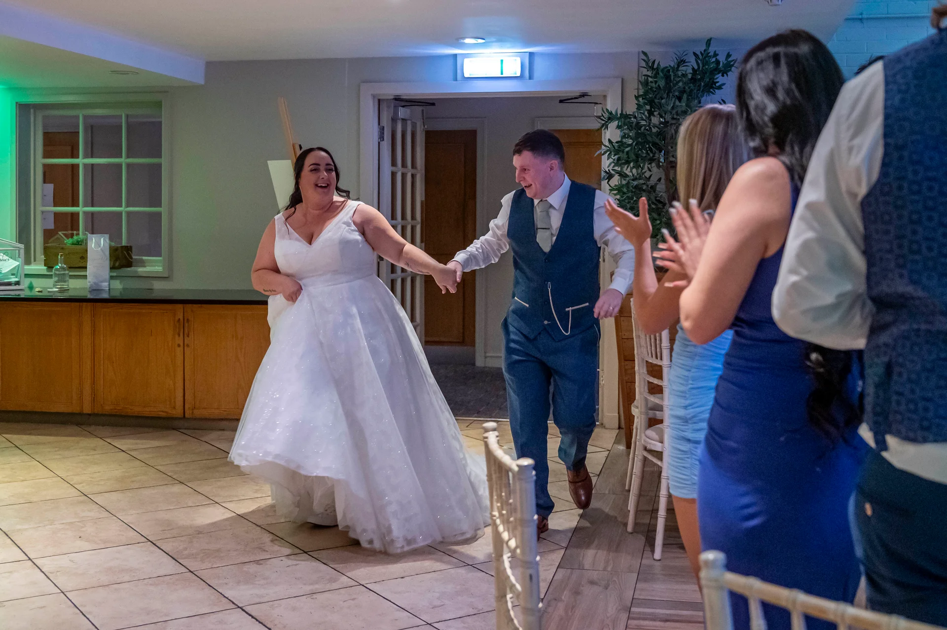 A newlywed couple excitedly enters the reception hand in hand, with the bride wearing a white a line wedding dress and the groom in a blue suit.