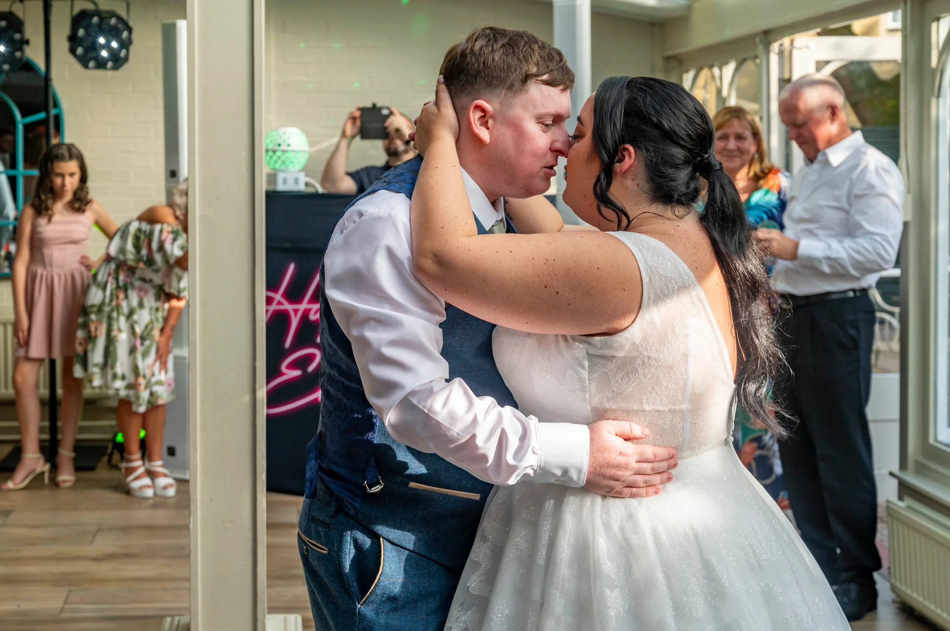 The newlywed couple shares a tender first dance together in a light-filled venue while guests watch on warmly from the background.