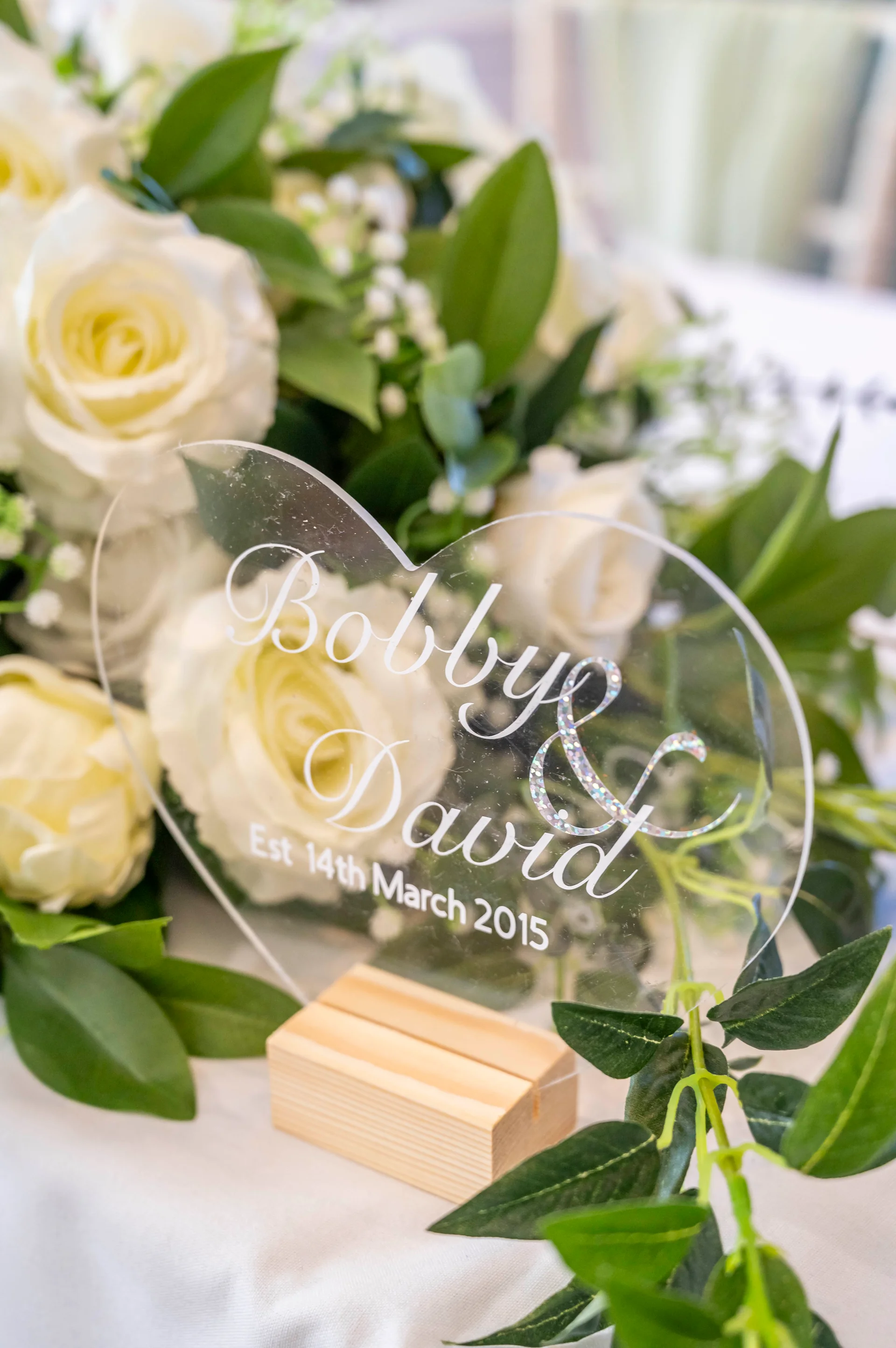A clear heart-shaped acrylic wedding sign with custom calligraphy sits nestled amongst a lush arrangement of white roses and green foliage.