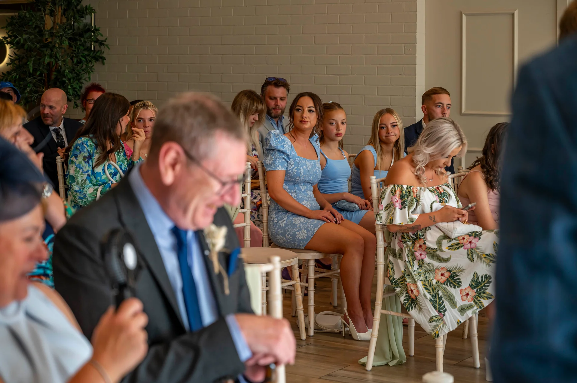 A candid shot of wedding guests seated indoors, dressed in smart casual attire and formal wear watching the ceremony unfold.