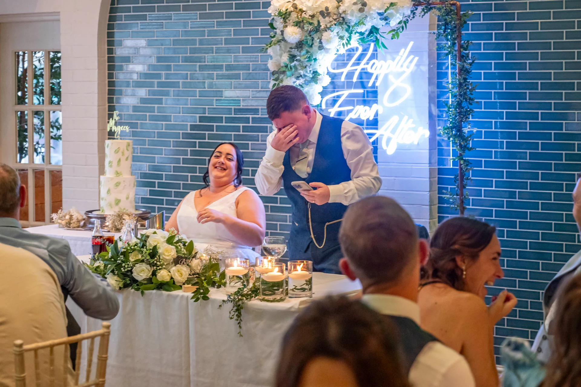 The groom covers his face while delivering an emotional speech next to the laughing bride at the top table, set against a glowing blue neon sign and a blue tiled wall.